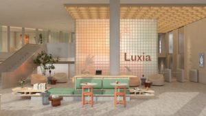 Interior view of the Luxia lobby featuring a backlit glass brick wall with a gradient of warm tones, soft seating areas, plants, and minimalist furniture creating a welcoming atmosphere.