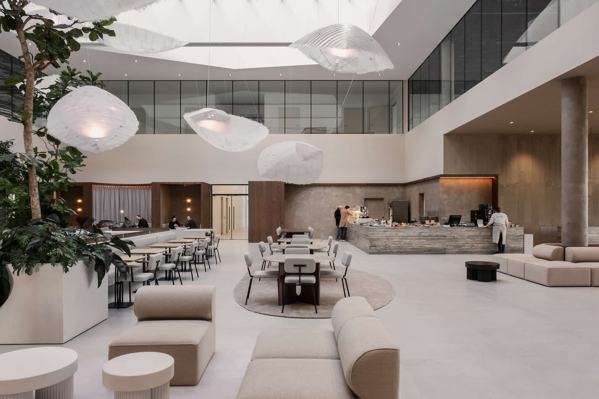Spacious and elegant cafeteria area with custom beige seating, round and rectangular tables, a central bar counter with stone finish, and sculptural pendant lights suspended from a skylight ceiling. Natural light enhances the contemporary interior design.