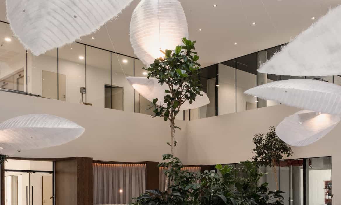Close-up of indoor trees and lush greenery under sculptural pendant lights in a modern atrium space, with glass-walled mezzanine offices visible above.