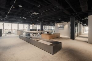 Open office space with custom-built seating platforms, high tables with wooden stools, and large windows offering panoramic city views. The design features exposed ceilings and integrated lighting for a contemporary atmosphere.