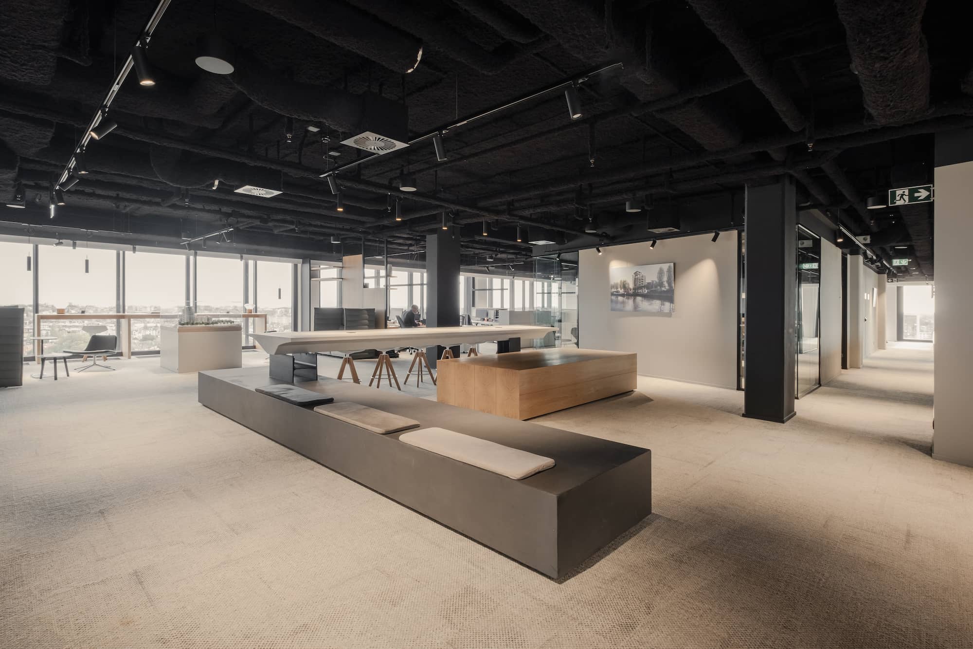Open office space with custom-built seating platforms, high tables with wooden stools, and large windows offering panoramic city views. The design features exposed ceilings and integrated lighting for a contemporary atmosphere.