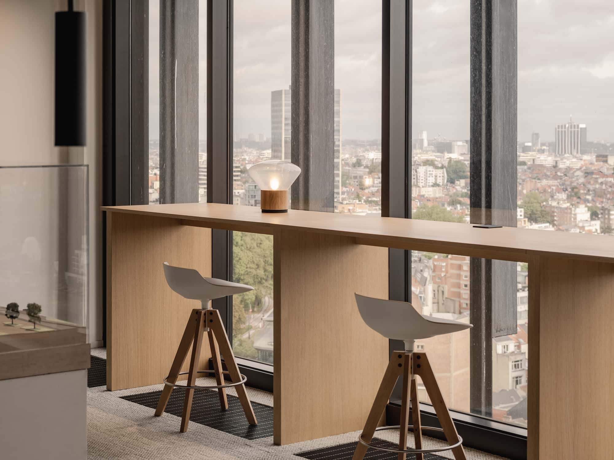 Tall wooden counter with minimalist bar stools overlooking the city through floor-to-ceiling windows, featuring a small table lamp and urban skyline in the background.