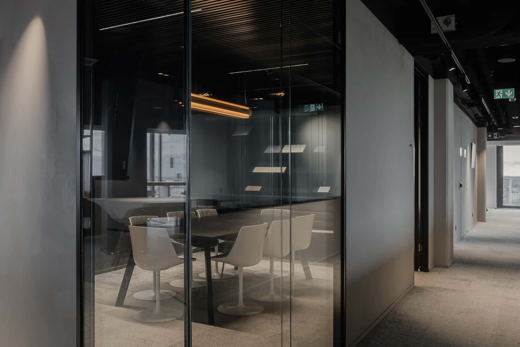 Glass-walled meeting room with a dark ceiling and modern white chairs, located along a softly lit office corridor.