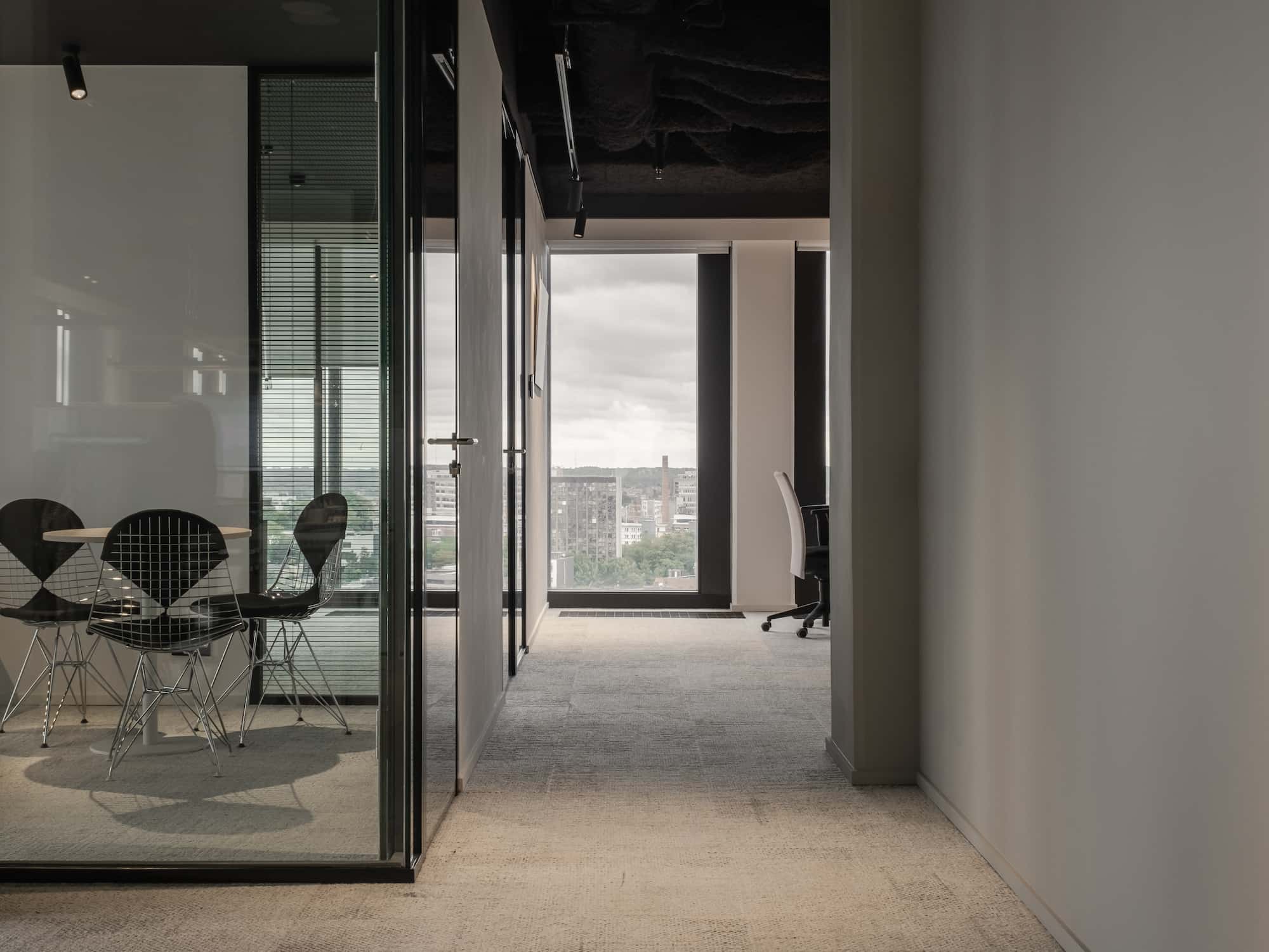 View of a glass-enclosed meeting room with black wireframe chairs and a round table, facing a large window with city views.