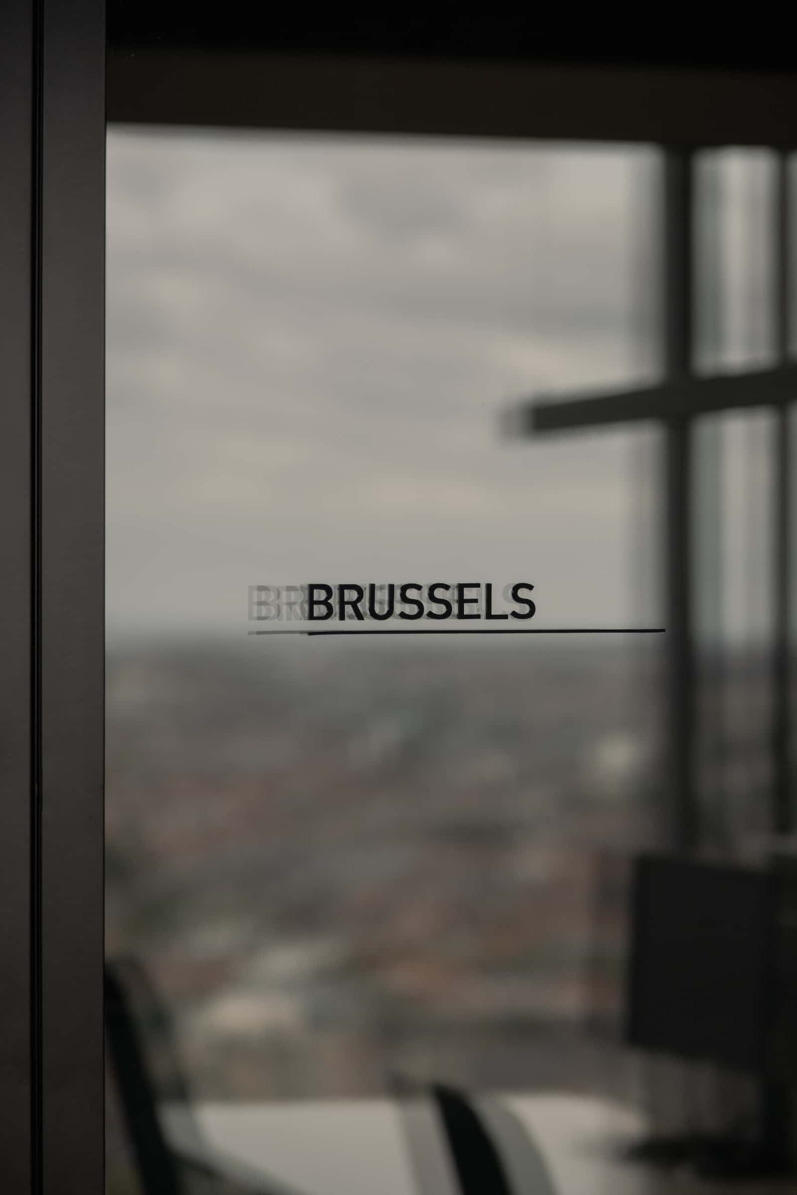 Close-up of a glass partition with the text 'BRUSSELS' printed on it, overlooking the city skyline through large windows.