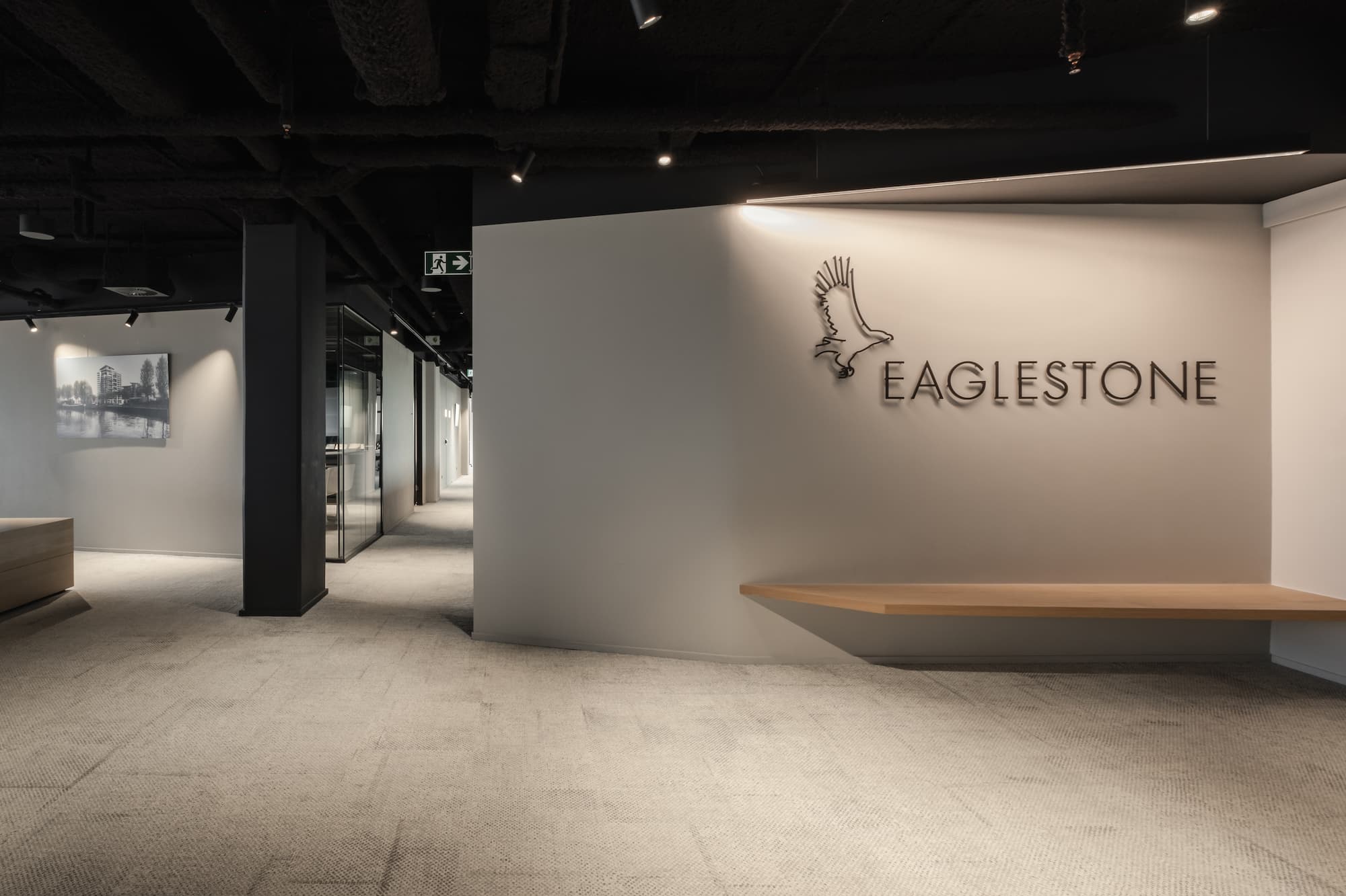 Reception area of Eaglestone offices featuring a minimalist floating wooden bench, a light grey wall with the Eaglestone logo and eagle graphic, and a corridor leading to glass-enclosed meeting rooms under a dark exposed ceiling.