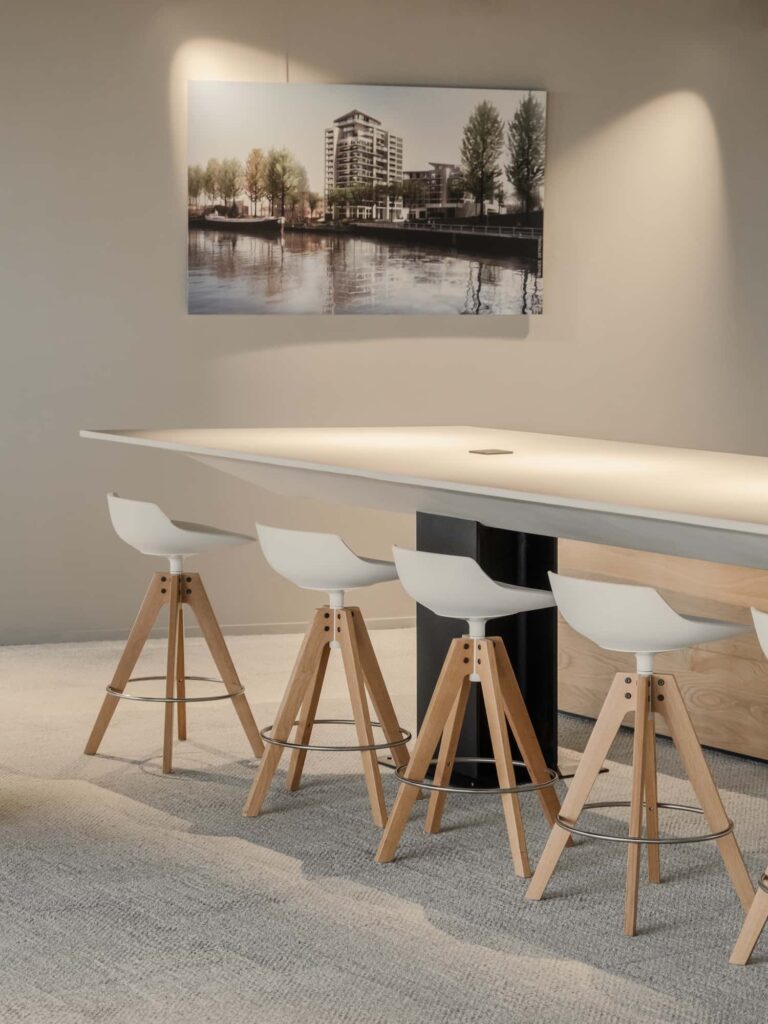 Minimalist high table with wooden stools in front of a wall-mounted architectural rendering of a modern residential building by the water.