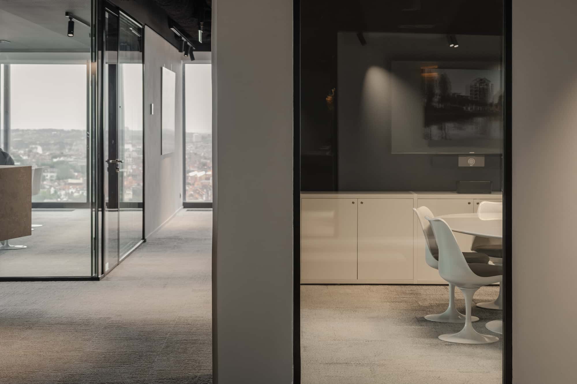 Interior view of a modern office corridor with glass-walled meeting rooms, featuring white Tulip chairs around a round table and panoramic city views in the background.