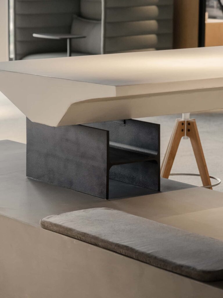 Close-up of a custom-designed central table with sculptural base in dark steel and integrated cushion seating in a modern office setting.