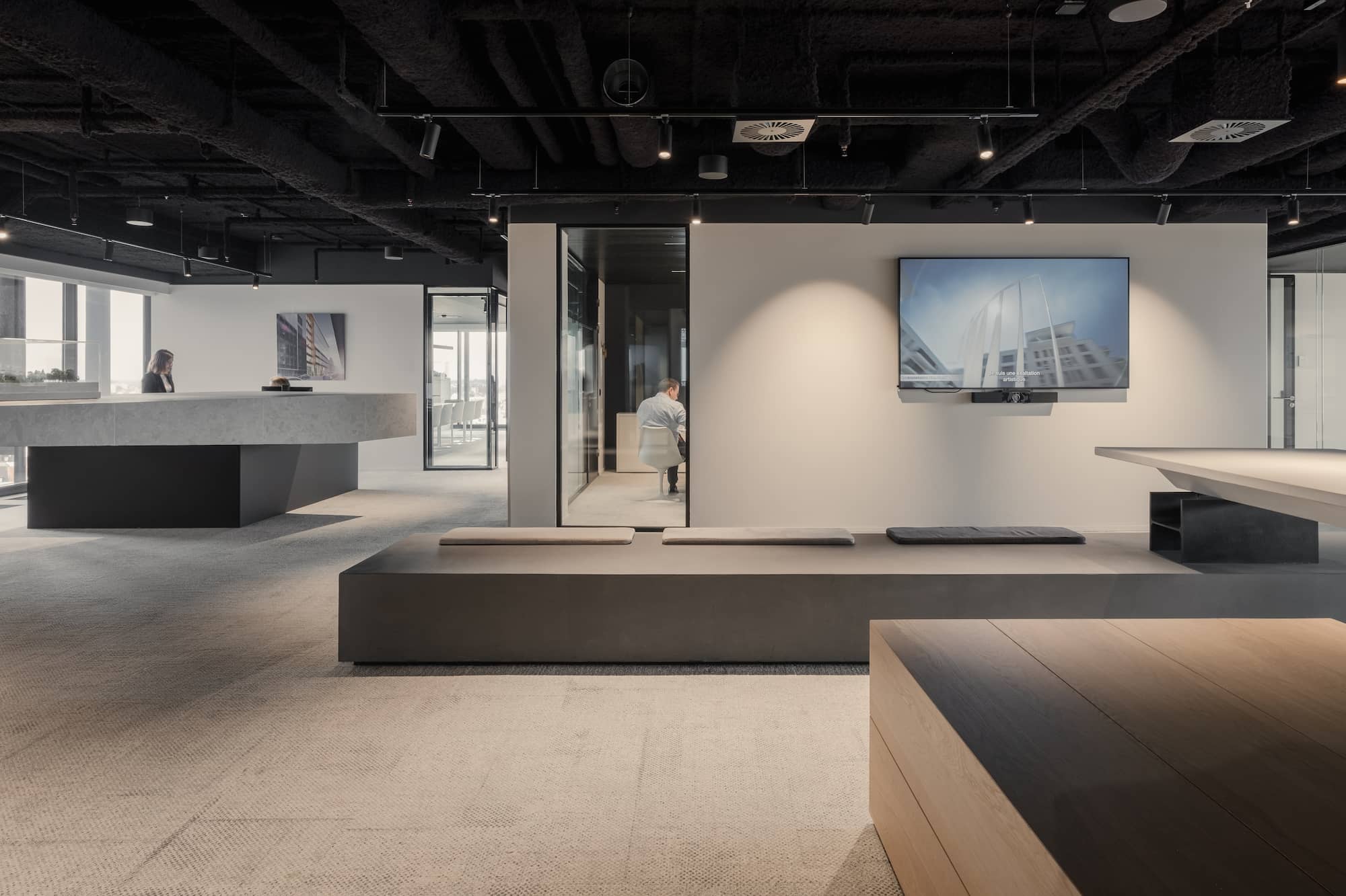 Modern office lounge area featuring built-in seating with grey cushions, a wall-mounted TV screen, and a sculptural concrete reception desk, all under a ceiling with exposed black acoustic treatment.