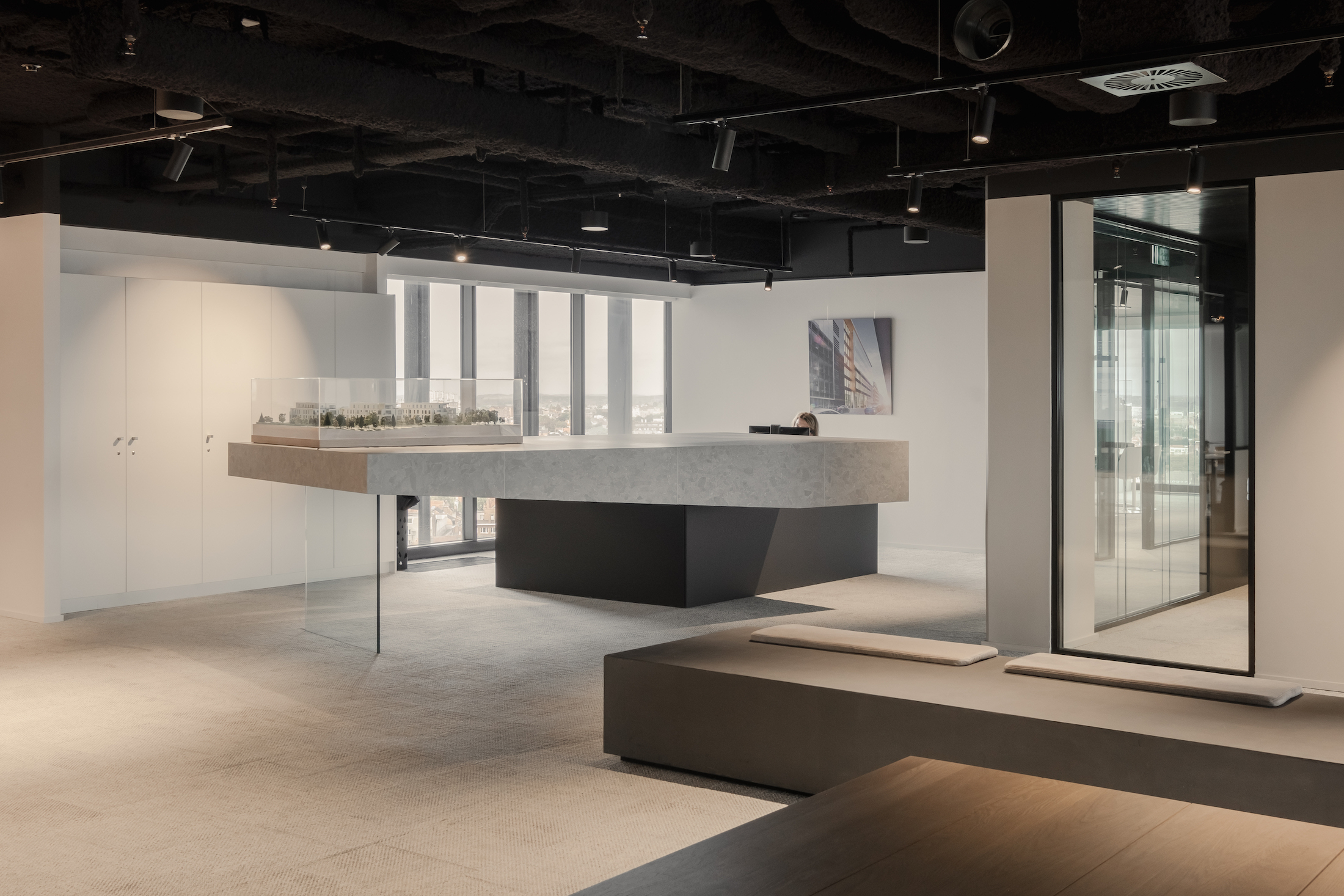 Minimalist office reception area with a sculptural concrete desk, architectural scale model under a glass case, and floor-to-ceiling windows providing panoramic city views.