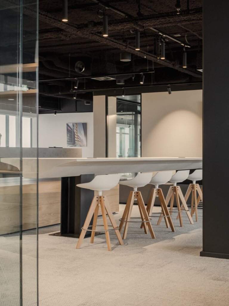 Detail view of a collaborative work area with high stools and a long central table, set under an exposed black ceiling with track lighting.
