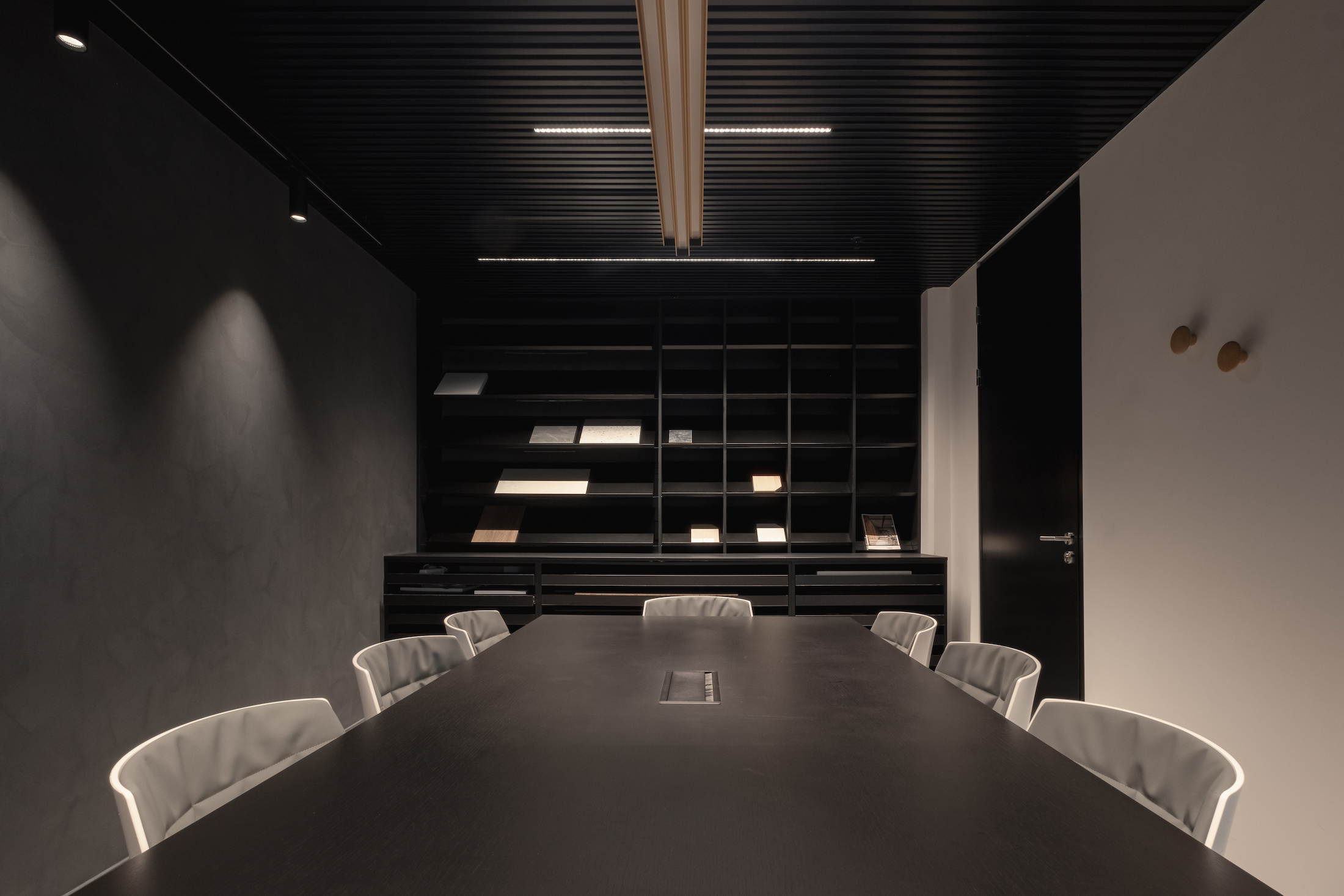 Interior view of a dark-toned meeting room featuring a black table, neutral chairs, and a back wall with material samples on open shelving.
