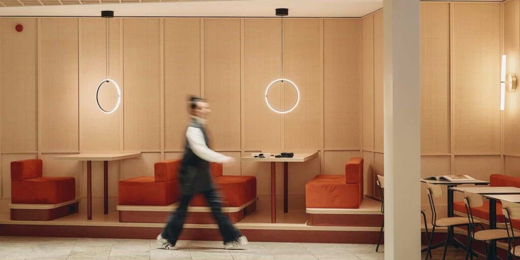 Interior view of a modern dining area with wooden panel walls, red cushioned booth seating, and circular pendant lights, as a person walks past in motion blur