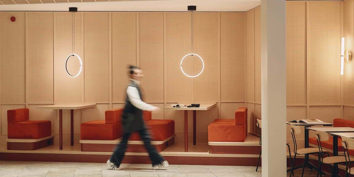 Interior view of a modern dining area with wooden panel walls, red cushioned booth seating, and circular pendant lights, as a person walks past in motion blur