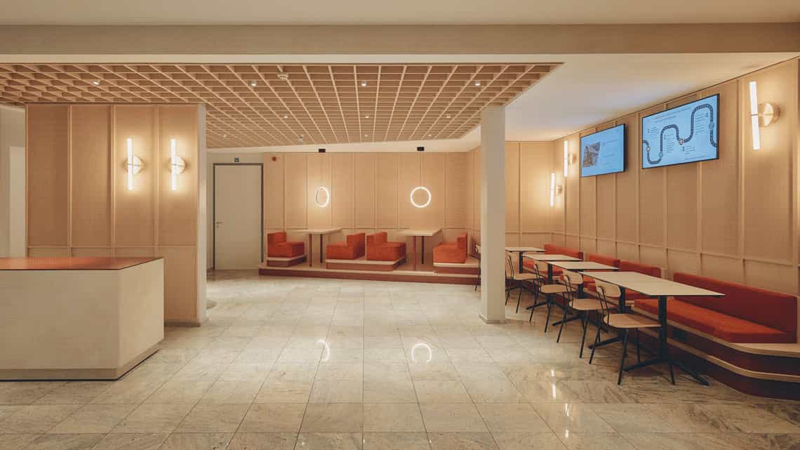 Wide-angle view of a bright dining area with integrated seating booths, red upholstered benches, wood paneling, and modern circular light fixtures on the wall and ceiling.