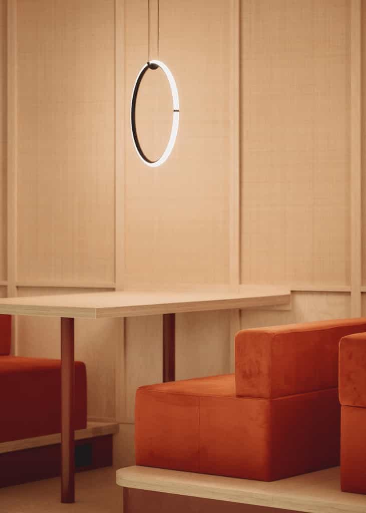 Close-up of a minimalist light ring fixture suspended above a light wooden table surrounded by red cube-like upholstered seats.