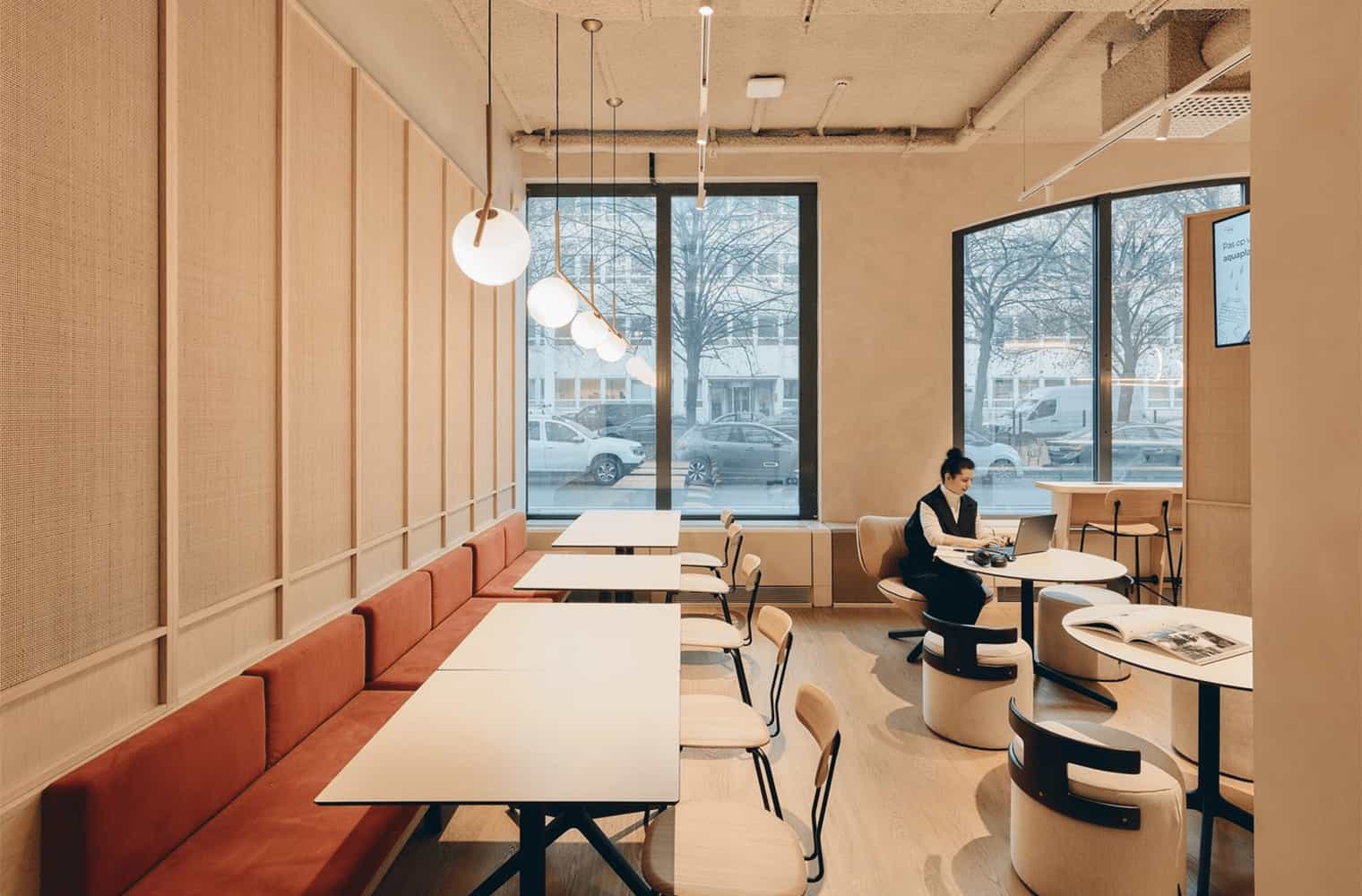 Dining space with long red-upholstered bench seating, white tables, and wooden chairs, adjacent to a large window and a separate work lounge area