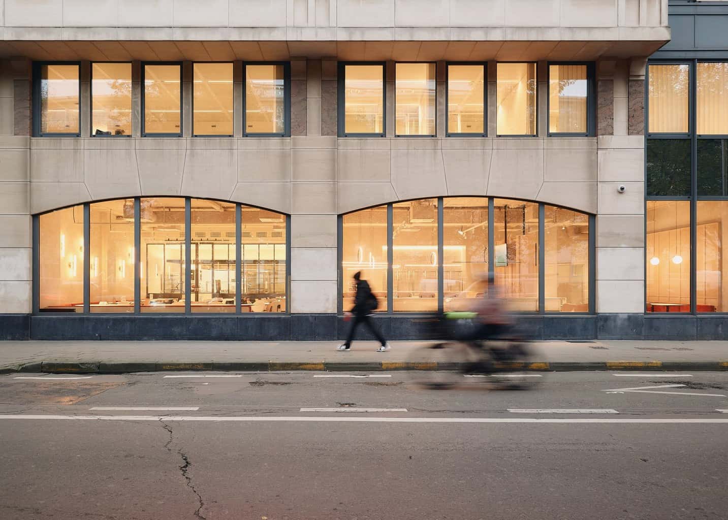 Street view of a building façade with large arched windows revealing a warmly lit interior dining space; pedestrians and a cyclist blur by in motion.