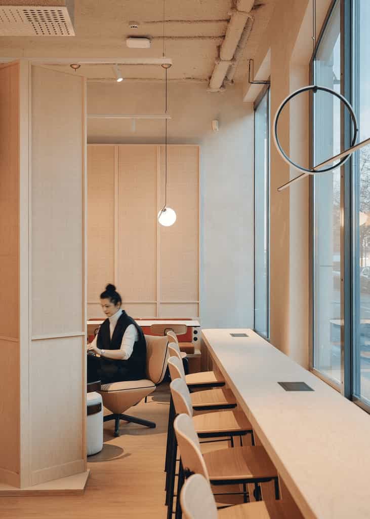 Narrow workspace with a tall bar-style counter along a large window, pendant lighting overhead, and a person working in a soft seating zone