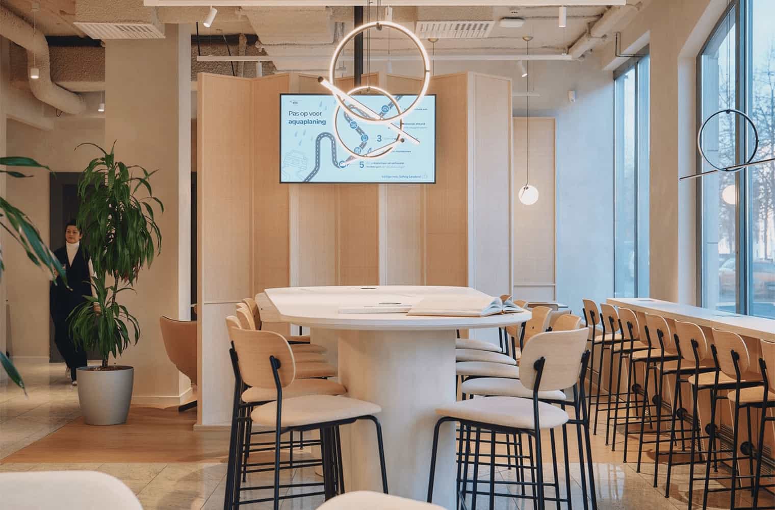 Central high table with wooden stools under a sculptural ring light, set against a backdrop of wood paneling and a digital screen.