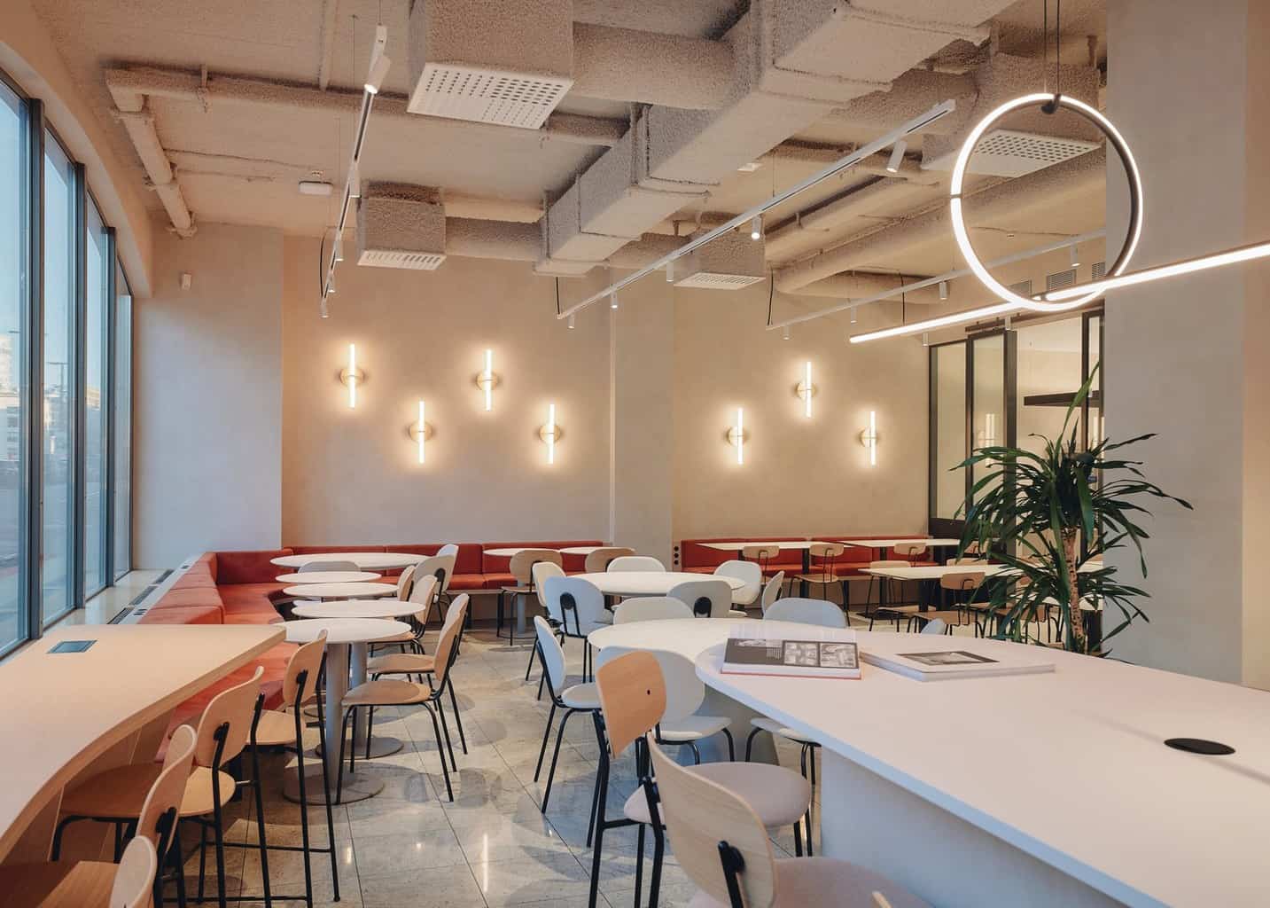 Wide-angle shot of a contemporary cafeteria with light-colored walls, red bench seating, pale wood chairs, and geometric lighting fixtures.