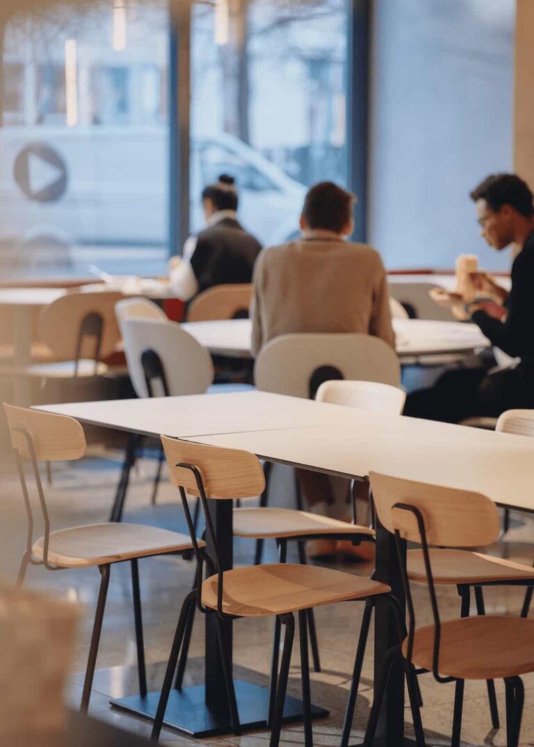 Group of people seated at minimalist white tables and wooden chairs, enjoying a meal in front of large windows.