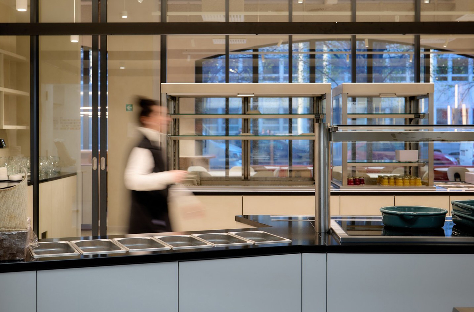 A woman moves behind a modern self-service counter, framed by glass walls and stainless-steel food displays