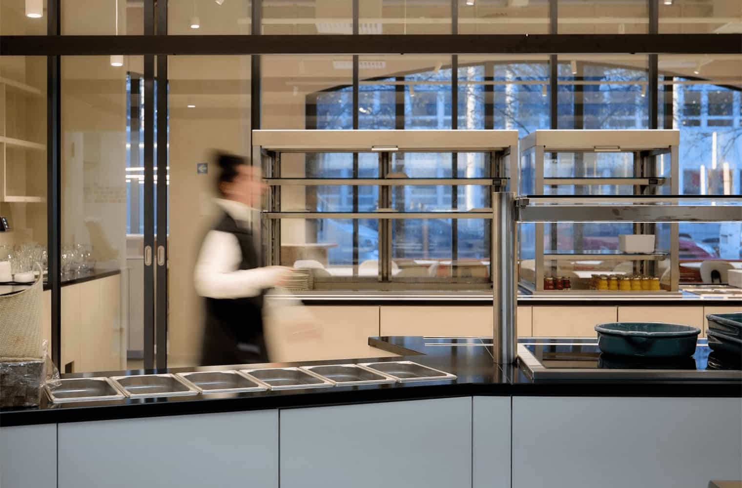 Self-service kitchen station with a blurred staff member in motion, stainless steel food displays and glass partition.