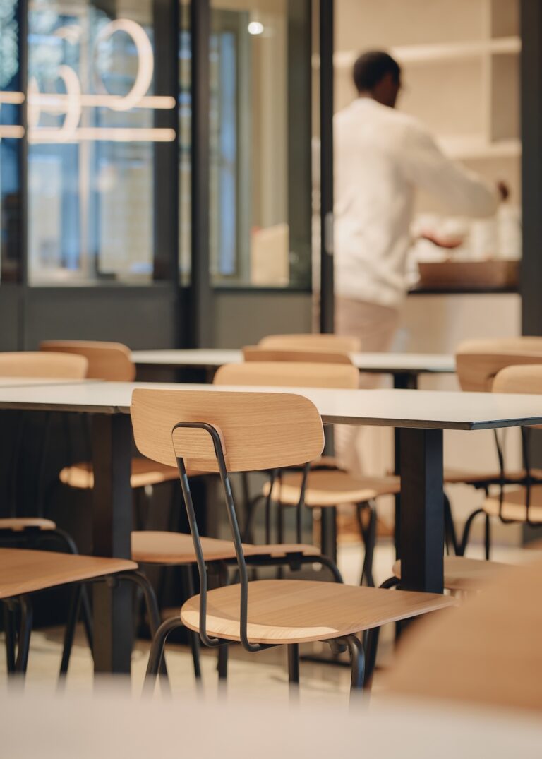 Close-up of dining area with wooden chairs and white tabletops, while a person prepares food in the background
