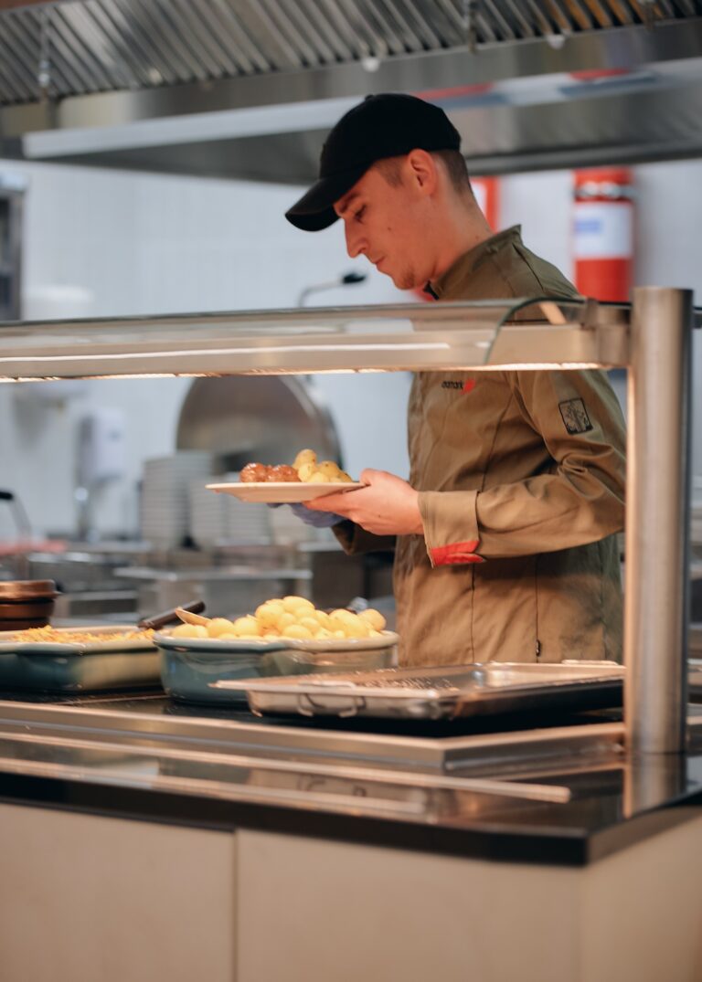 A chef wearing a cap and uniform serves a plate of potatoes and meatballs in the kitchen's hot food station