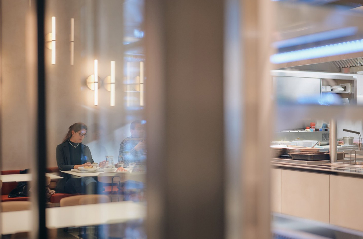 View through a glass partition showing a woman eating lunch under warm lighting, with a glimpse of the kitchen in the background.