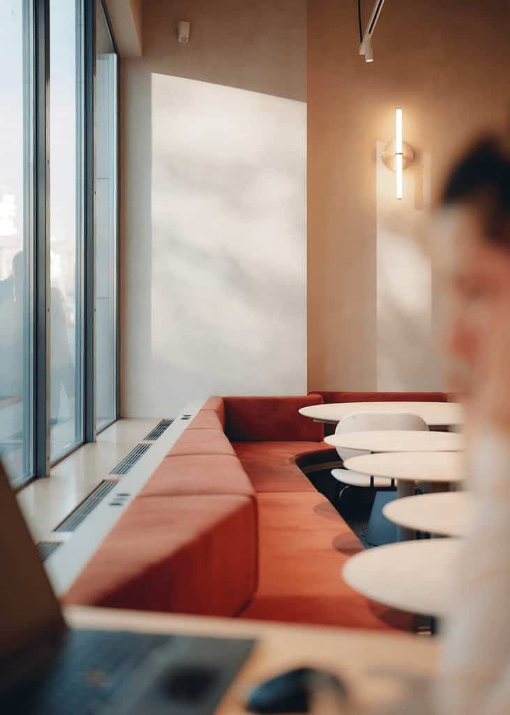 Close-up of a corner seating area by tall windows, featuring red upholstered benches and small white round tables, softly lit by daylight.