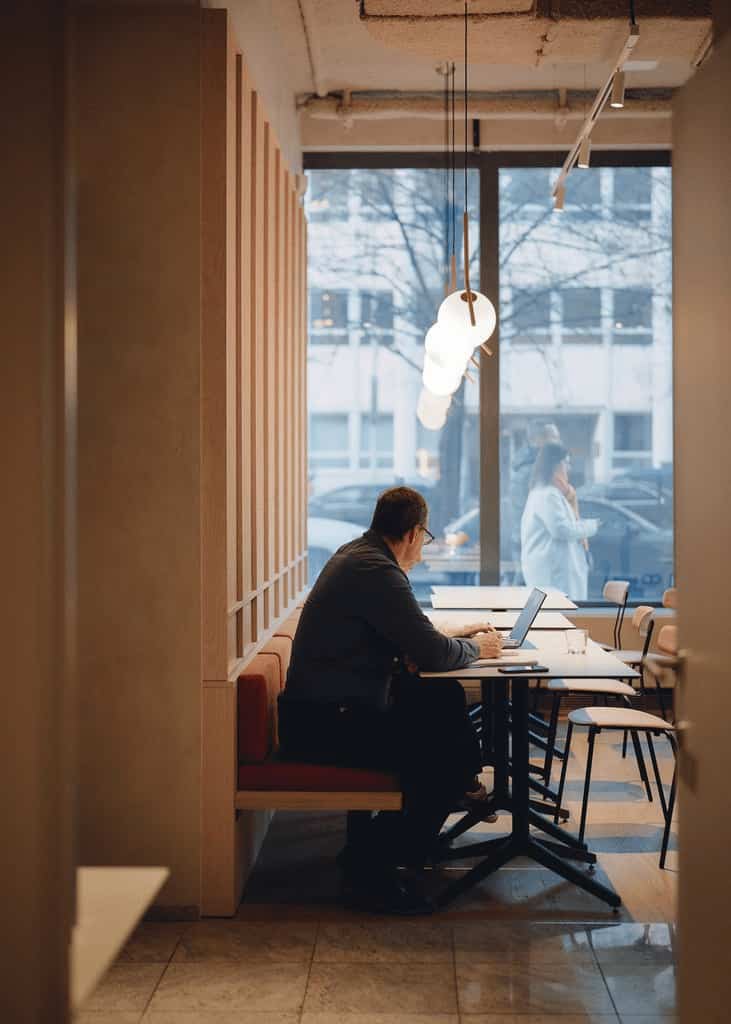 Cozy corner in a dining area with a man working on a laptop at a table near a large window, under warm pendant lights and a soft ambient glow