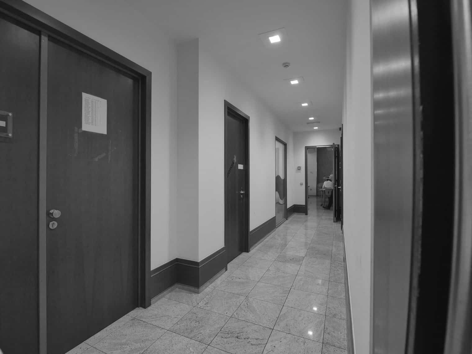 Black and white image of a plain office hallway with closed wooden doors and tiled flooring