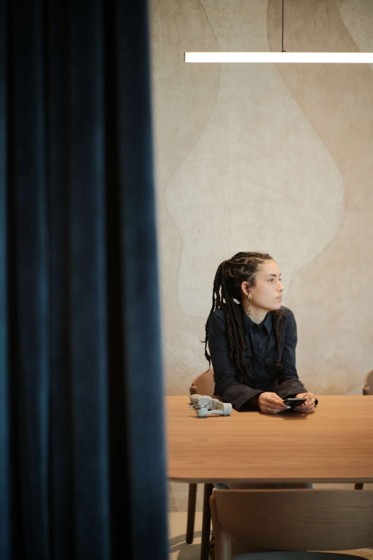 Woman seated at a wooden table in a minimalist meeting space with sculptural wall texture and pendant light.