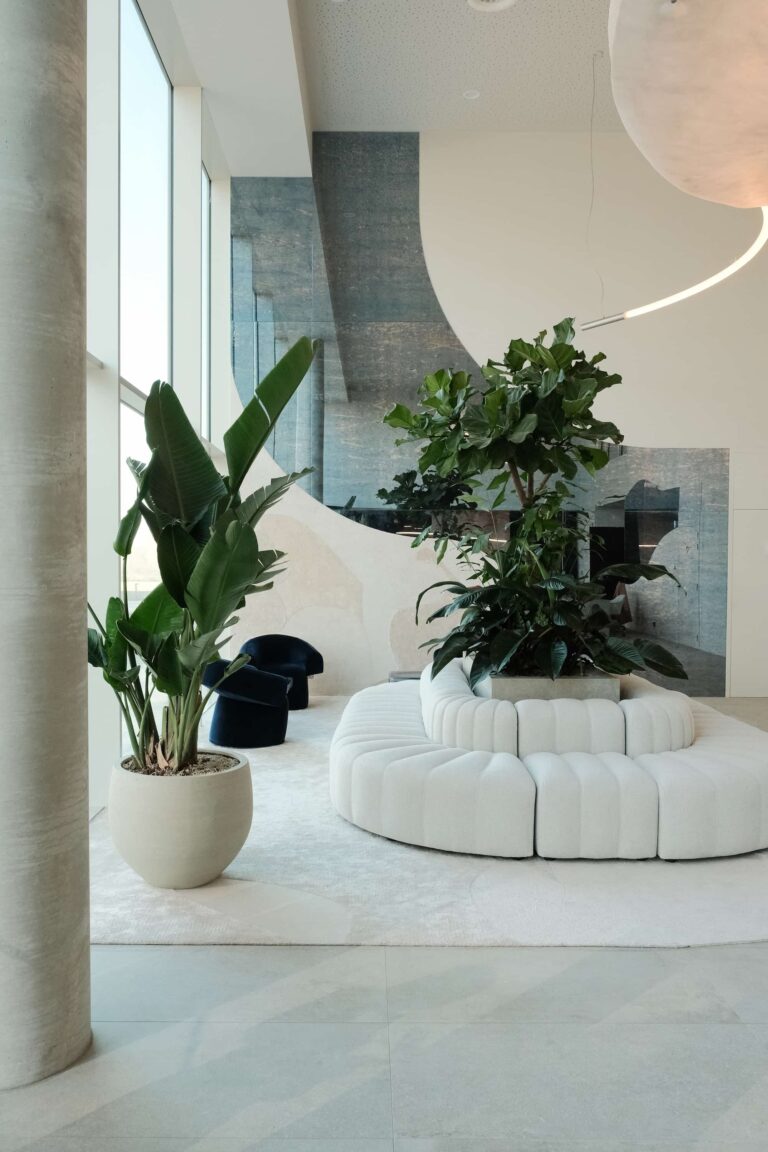 Interior lounge area with curved white modular seating, indoor plants, and natural light from floor-to-ceiling windows