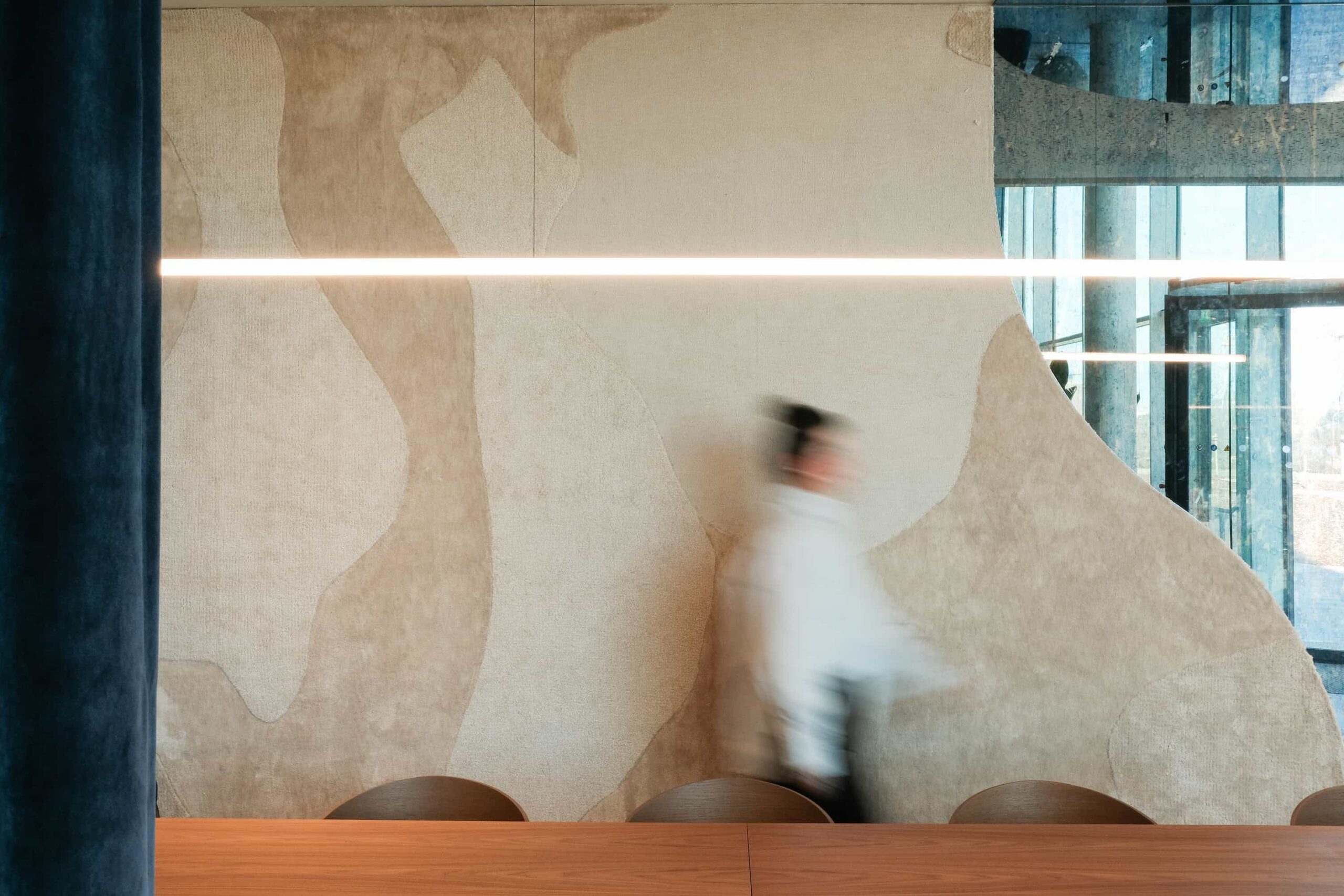Blurred woman walking past an abstract sculptural wall with linear pendant light above a wooden meeting table