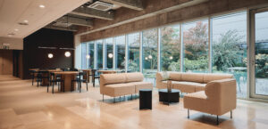 Modern lounge and dining area with beige sofas, wooden tables, and large windows overlooking a garden with greenery and outdoor seating