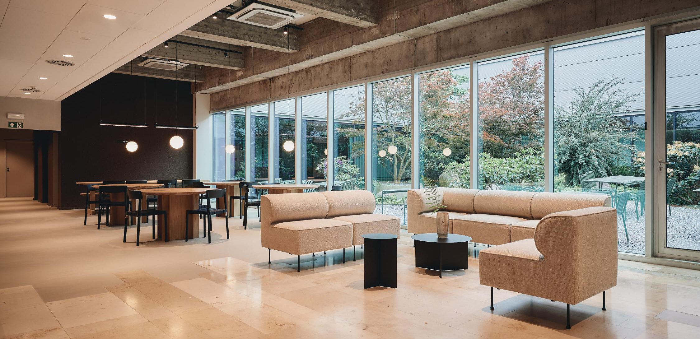Modern lounge and dining area with beige sofas, wooden tables, and large windows overlooking a garden with greenery and outdoor seating