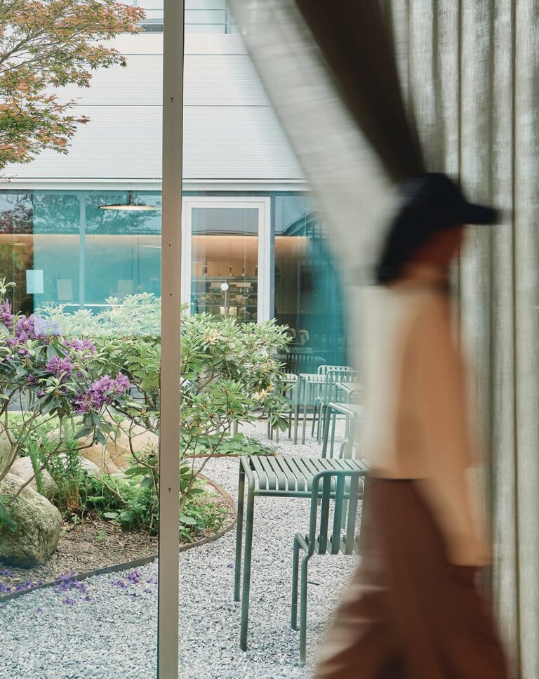 View of a tranquil garden with green chairs and blooming plants seen through a glass window with sheer curtains, as a blurred person walks by inside