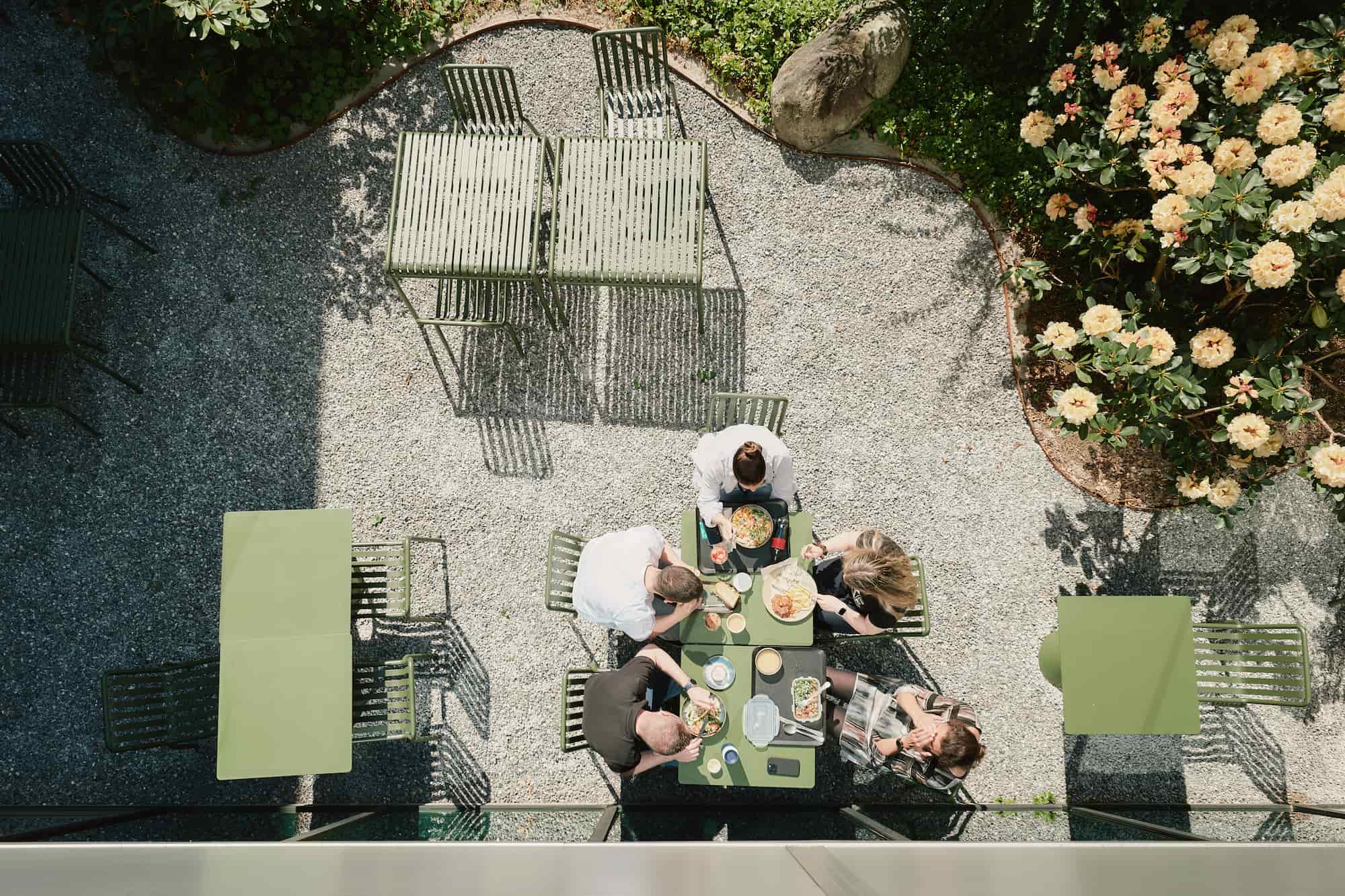 Top-down view of five people having lunch together at a green outdoor table in a garden setting with gravel ground, flowers, and greenery