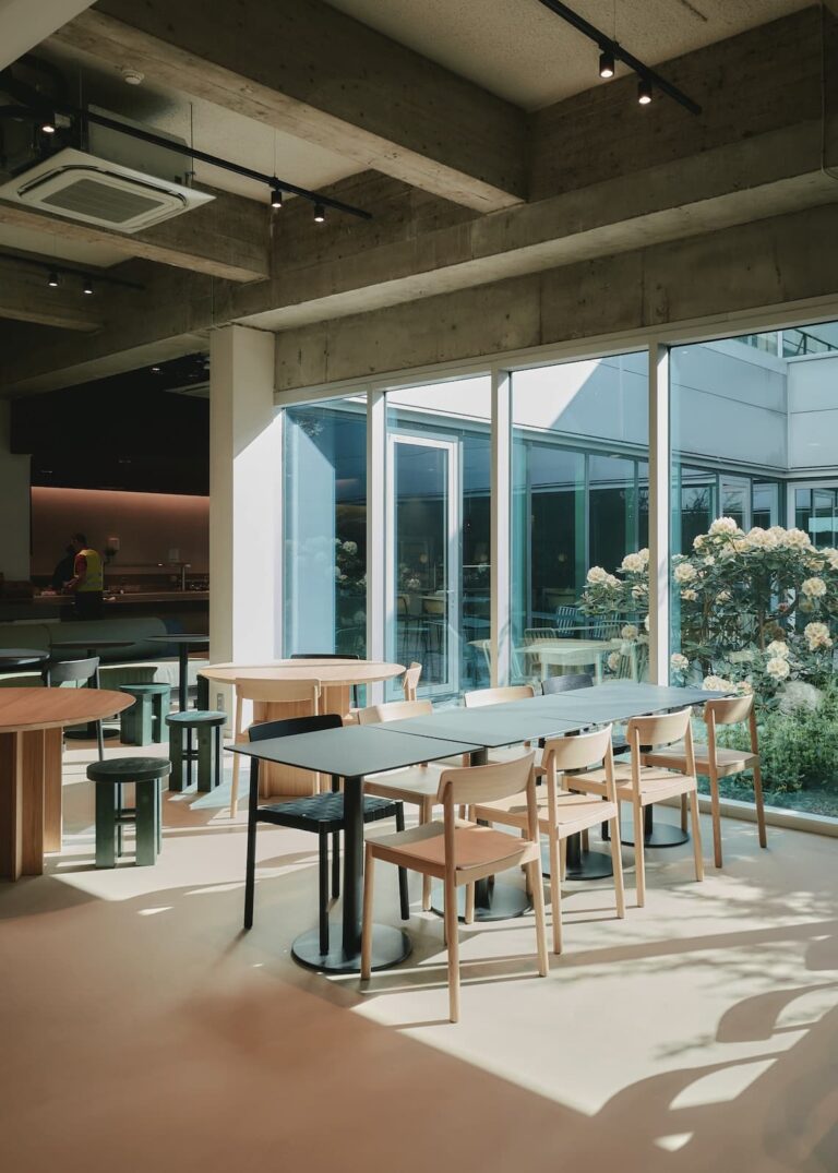 Bright dining area with wooden tables and chairs next to large glass windows overlooking a lush courtyard garden