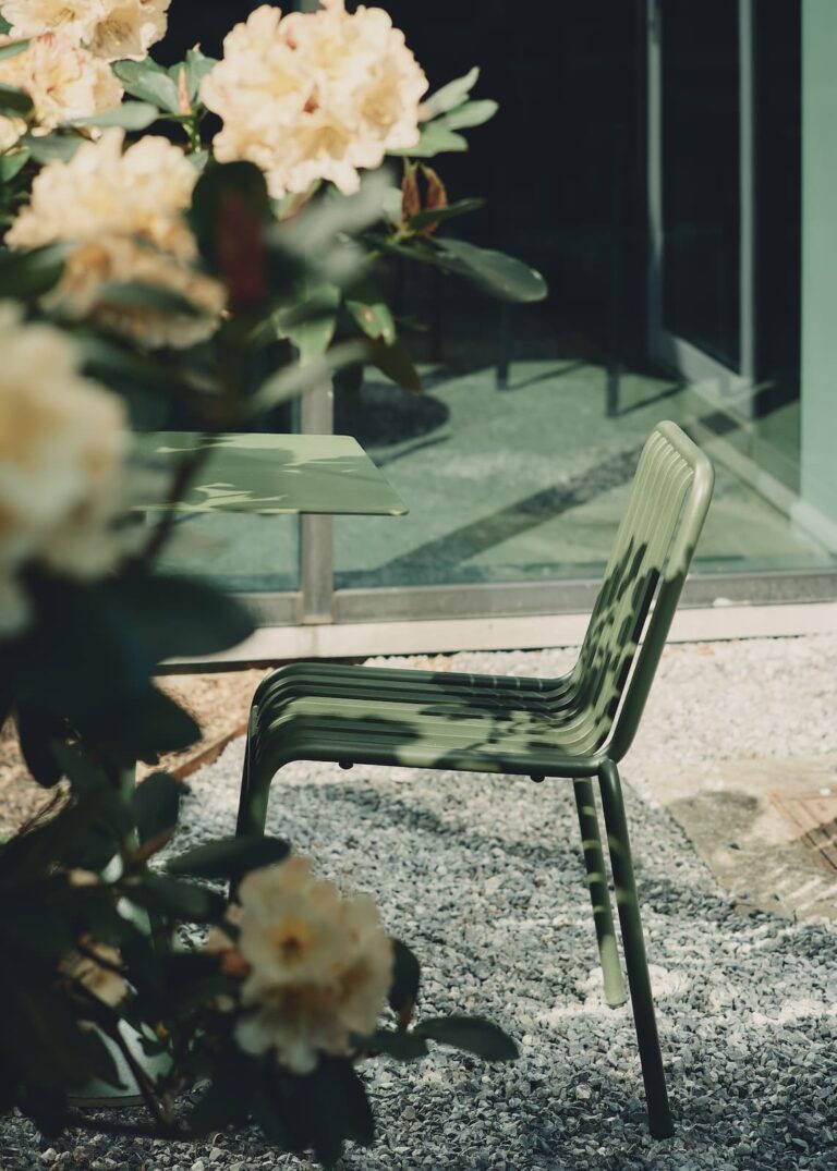 Close-up of a green metal outdoor chair next to a table, surrounded by blooming flowers and casting dappled shadows on gravel