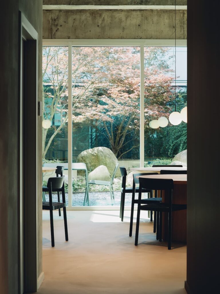 Interior view from a corridor toward a shared dining space with black chairs and large windows opening to a garden with trees and boulders