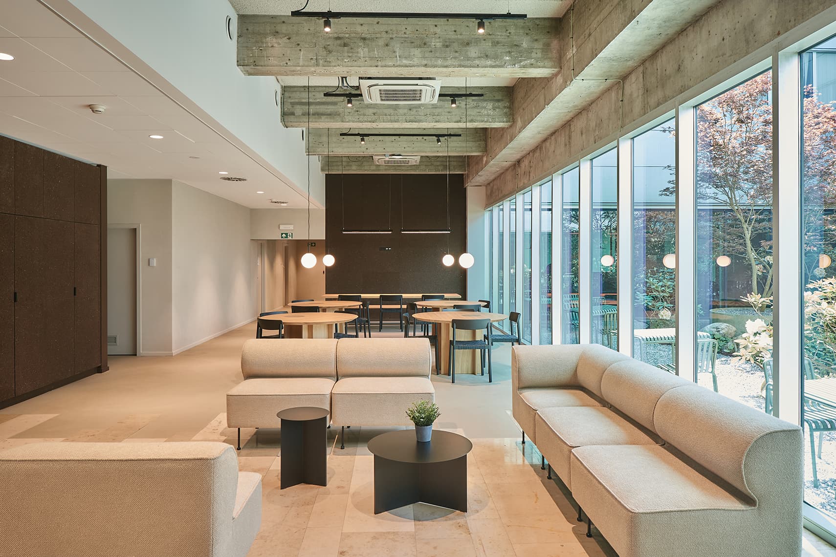 Bright lounge area with beige sofas, black round coffee tables, and floor-to-ceiling windows overlooking a garden