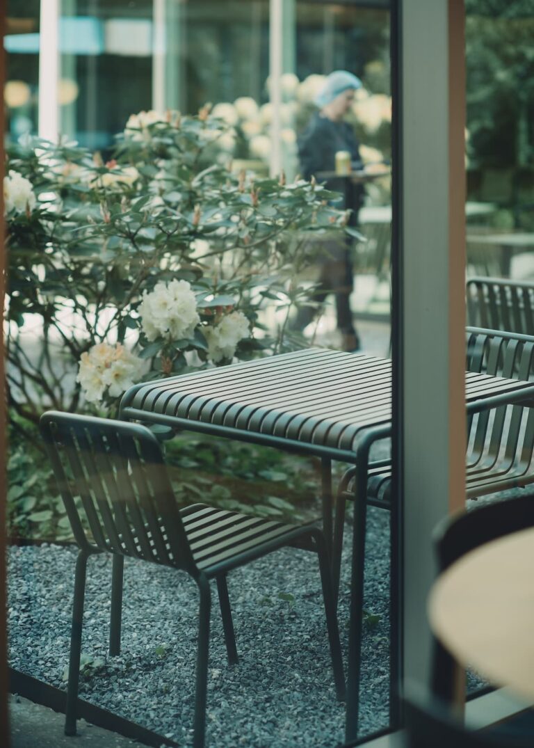 Outdoor dining area seen through a window, with striped green chairs and blooming plants