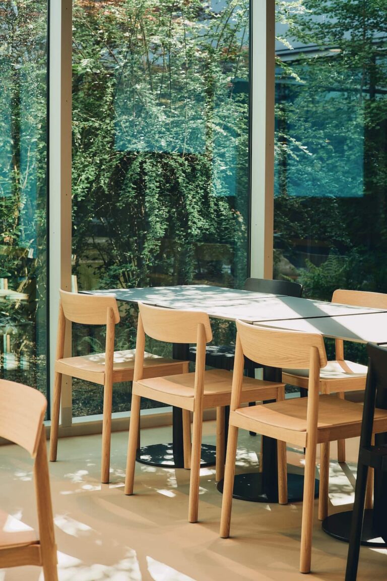 Light wooden chairs around a rectangular table next to large windows reflecting lush green vegetation
