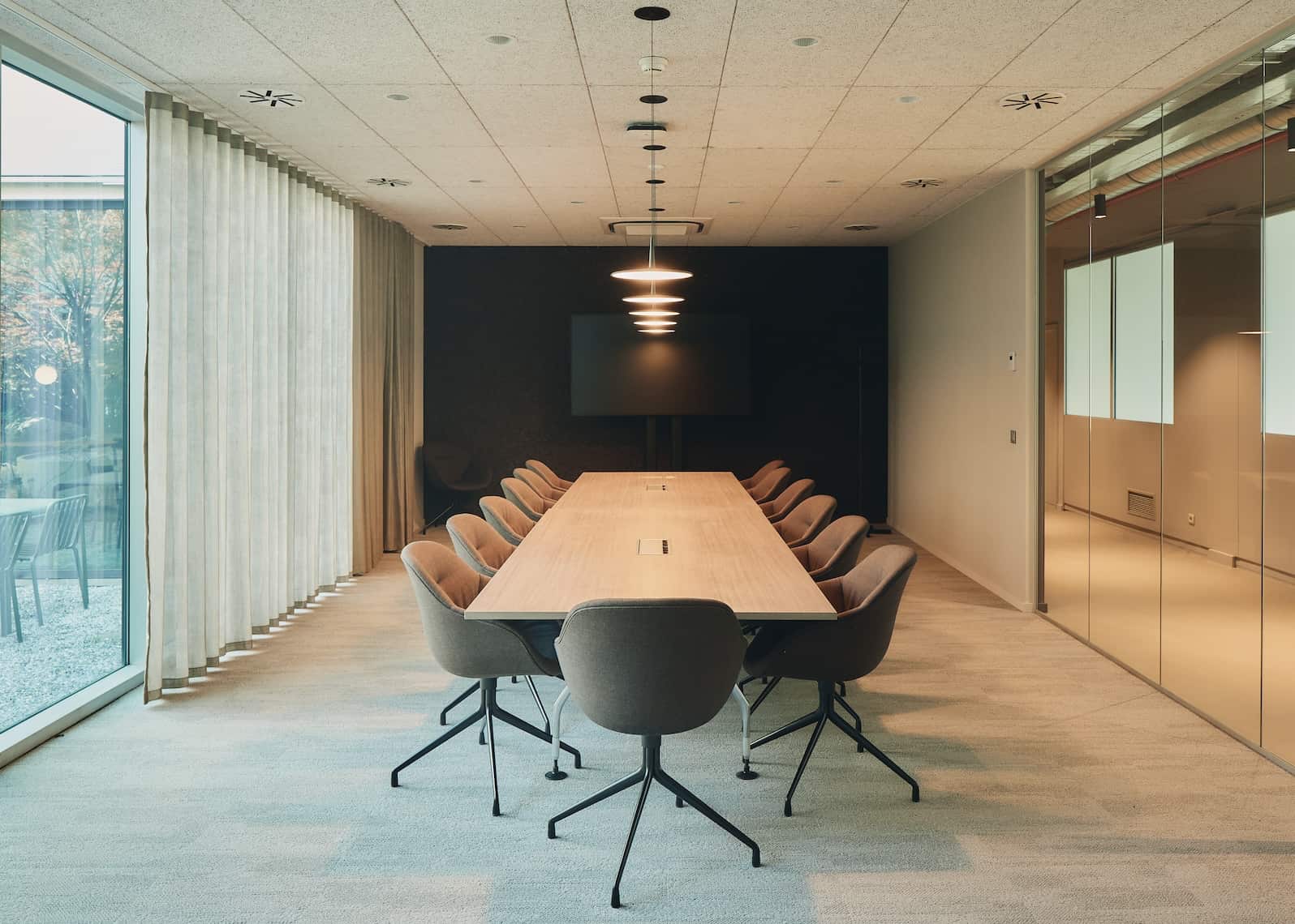 Modern meeting room with a long wooden conference table, brown swivel chairs, and large windows with curtains offering natural light