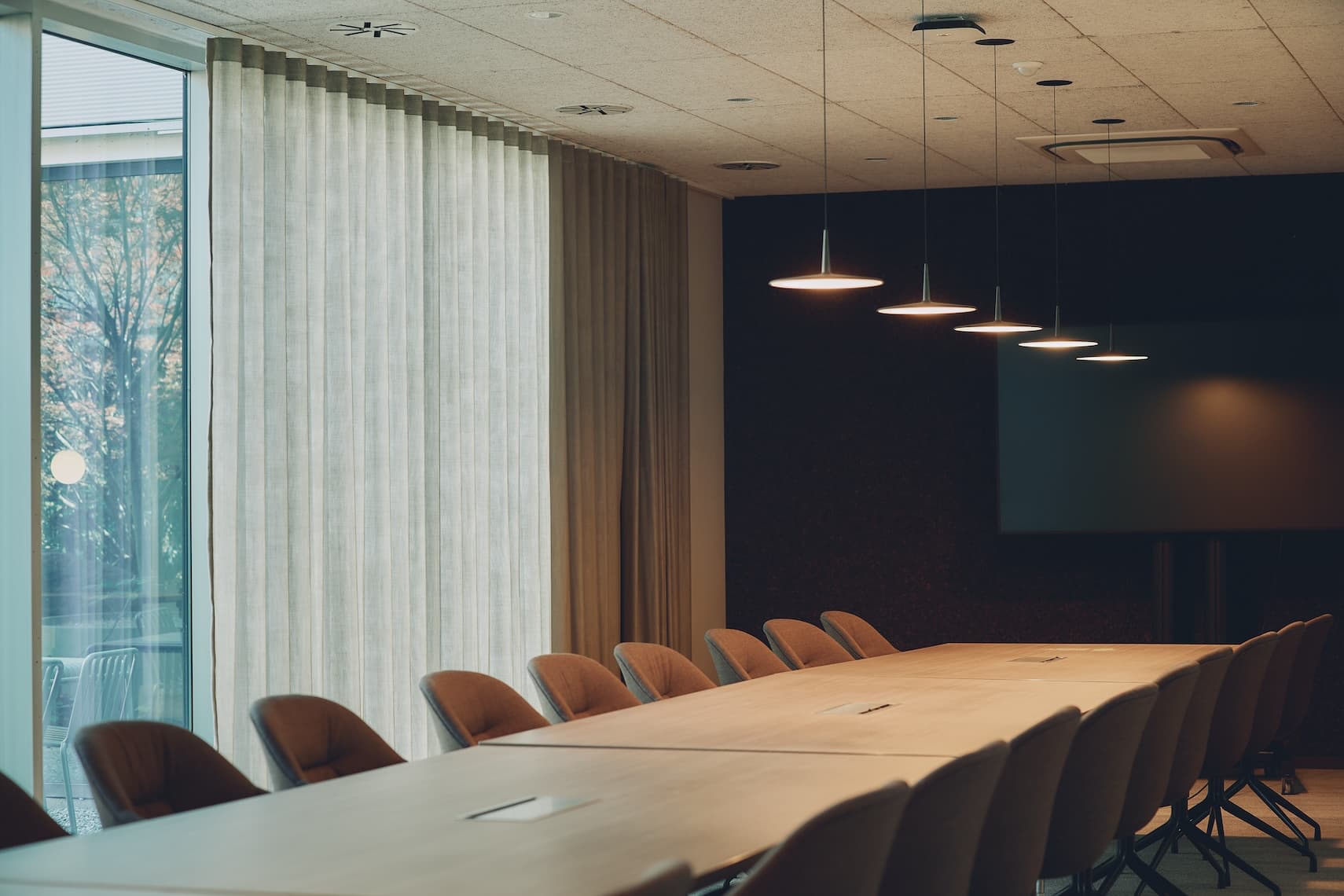 Contemporary meeting room with a long wooden table, brown swivel chairs, and a row of minimalist pendant lights above